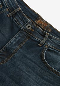 Tamsiai mėlyni džinsai su oranžinėmis siūlėmis, metalinėmis mygtukų uždarymo systemomis ir etikete "Premium Denim Goods" esančia šalia juosmens.