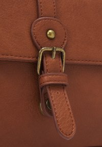 Szczegół brązowej skórzanej torby z mosiężną klamrą, paskiem i przeszyciami. Powierzchnia ma teksturowane wykończenie z wzmocnionymi elementami metalowymi.