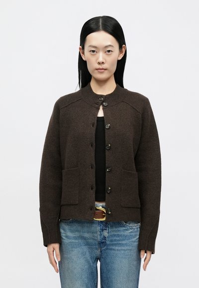 Pruun villane kardigan ümmarguse kaelusega, nööbitav eest ja kahe esitaskuga. Stiilitud musta topi ja hele siniste teksadega.