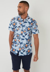 Chemise à manches courtes, bleu clair, avec un motif floral comprenant des fleurs orange et des feuilles foncées. Portée avec un short marine, le tissu semble lisse et léger.