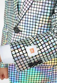 Kareline metallik ülikond, millel on ruudukujuline muster iridescentsetes värvides. Mustad nööbid ja valge sildiga "Oppo Suits" varrukal.