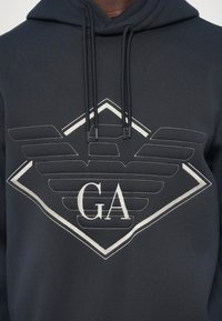 Tume kapuutsiga sviiter, millel on rindkeres suur tikanditud stiliseeritud kulli logo ja "GA" initsiaalid, kapuutsi küljes rippuvad nöörid.