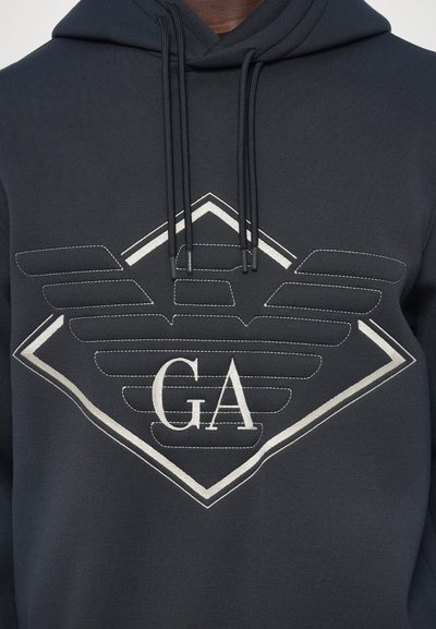 Temna kapuca z velikim vezenim stiliziranim logotipom orla in inicialkami "GA" na prsih, vrvici, ki visijo iz kapuce.