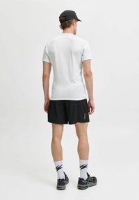 Mans sportiga kläder består av en figurnära vit t-shirt med kort ärm, svarta shorts och svarta sneakers med vita mönstrade strumpor.