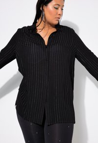 Schwarzes Hemd mit silbernen vertikalen Streifen, langen Ärmeln, Kragen und entspannter Passform, kombiniert mit schwarzen Leggings mit kleinen Verzierungen.