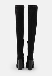 Bottes noires cuissardes en daim souple avec un talon haut épais et des fermetures éclair sur les côtés. Texture lisse avec des détails minimaux.