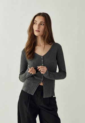 Kobieta z długimi brązowymi włosami, ubrana w szary, prążkowany, zapinany na guziki kardigan oraz czarne spodnie, patrząca na bok na tle gładkiego tła.