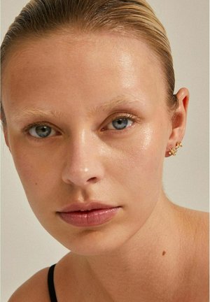 Gros plan sur un visage à la peau claire, au teint texturé, avec des yeux bleus et un maquillage minimal. Porte un ear cuff en or avec des accents floraux.