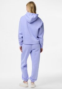 Kvinne står med ryggen til iført en lilla hettegenser og matchende joggebukser med hvite joggesko foran en hvit bakgrunn.