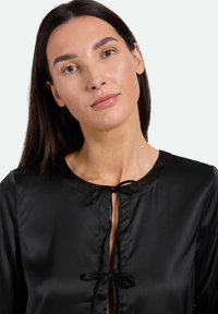 Schwarze Satinbluse mit rundem Ausschnitt, einer Schleifenverschluss vorne und langen Ärmeln. Glatte Textur mit minimalem Glanz.