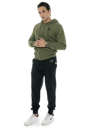 Un hombre luce una sudadera con capucha y cremallera de color verde oliva, pantalones deportivos negros y zapatillas blancas sobre un fondo blanco liso.