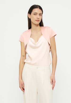 Γυναίκα με μακριά σκούρα μαλλιά, που φοράει γαλάζιο σατέν τοπ με γιακά cowl, ανοιχτό ροζ κοντό σακάκι και λευκό ψηλόμεσο παντελόνι, στέκεται μπροστά σε λευκό φόντο.