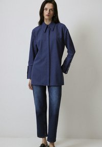 Navy blue striped long-sleeve shirt with a pointed collar, featuring buttoned cuffs, paired with straight-leg dark denim jeans.