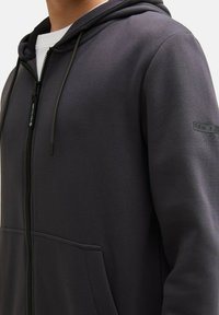 Ingrandimento di una persona che indossa un felpa con zip di un grigio scuro, dotato di tasche frontali e un piccolo logo sulla manica superiore.