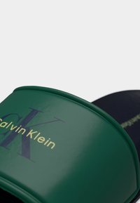 Κοντινή λήψη ενός πράσινου σανδαλιού τύπου σλάιντ με σήμανση Calvin Klein και μαύρη επιφάνεια για το πόδι σε ανοιχτό φόντο.