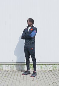 Fekete sportos melegítő szett narancssárga részletekkel, cipzáros dzseki, hozzáillő nadrág, és fekete sportcipő piros talppal. Egyszerű háttér, álló póz.