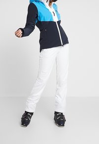 Lyžařská bunda v námořnické modři a světle modré barvě s bílým předním zipem, kombinovaná s bílými lyžařskými kalhotami a černými lyžařskými botami s barevnými akcenty.