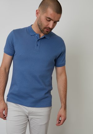 Polo bleu avec un motif texturé, manches courtes et patte de boutonnage à deux boutons, associé à un pantalon de couleur claire.