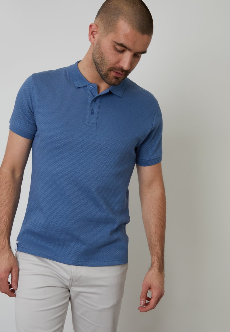 Modrá polo košile s texturovaným vzorem, krátké rukávy a dvoubarevný límeček, v kombinaci se světle zbarvenými kalhotami.