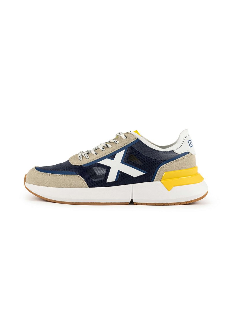 Textured navy fabric sneaker with beige suede accents, white branding, thick white midsole, yellow heel detail, and grey laces.