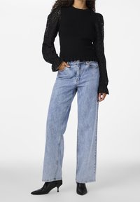 Musta neulepaita teksturoitujen hihojen kanssa, vaaleat denimpussihousut ja teräväkärkiset mustat korkokengät. Paidassa on röyhelöhelma ja kuvioita.