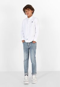 Weißes Hemd mit Knopfleiste und kleinem Logo auf der Brust, kombiniert mit hellblauen Jeans und grauen Turnschuhen. Glatte Stoffqualität und klassischer Schnitt.