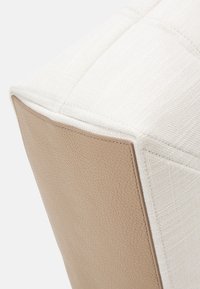 Vierkante poef met een gestructureerde witte stof aan de bovenkant en een gladde beige leren zijkant. Stiksel is zichtbaar langs de randen.