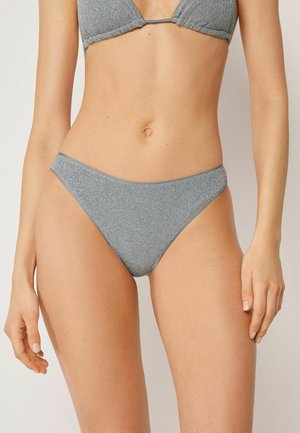 Kobieta ubrana w srebrnoszare błyszczące bikini – dół i pasujący top, stojąca na jednolitym jasnym tle.