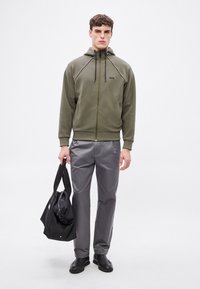 Zielona oliwkowa bluza z kapturem zapinana na zamek, z czarnymi akcentami, szare spodnie i czarna torba. Gładka tekstura, luźny krój i kaptur ze sznurkiem.