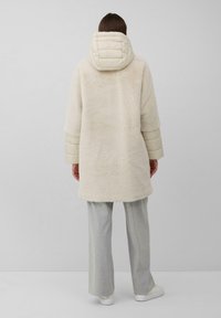 Crème-kleurige, fuzzy jas met een puffy capuchon en geborduurde mouwen. Zachte textuur, oversized ontwerp, valt tot halverwege de dij. Gecombineerd met lichtgrijze broeken.