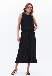 Schwarzes ärmelloses Maxikleid mit einem strukturierten Wellenmuster, das einen lässigen Schnitt und einen fließenden Saum hat, kombiniert mit schwarzen Riemchenhighheels.