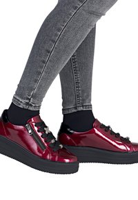 Glanzende rode plateau sneakers met zwarte veters en ritssluiting, gedragen met zwarte sokken en grijze skinny jeans.