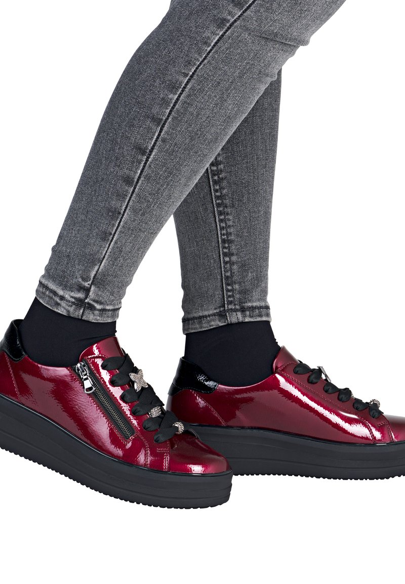 Glanzende rode plateau sneakers met zwarte veters en ritssluiting, gedragen met zwarte sokken en grijze skinny jeans.