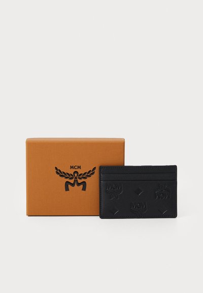 Svart lær kortholder med preget MCM-logo og diamantdetaljer, vist ved siden av en oransje gaveeske med merket logo.
