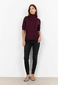Burgundi színű, rövid ujjú garbó pulóver, amely fekete skinny farmernadrággal és mintás szandálokkal van párosítva. Puha tapintású, testhez simuló dizájn, minimális részletekkel.