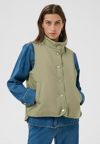 Ärmellose Weste in Olivgrün mit hohem Kragen, Druckknopfverschluss und Vordertaschen, kombiniert mit einem blauen Denim-Hemd mit langen Ärmeln.