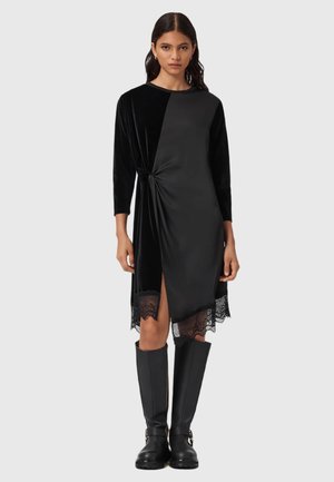 Zwart fluwelen jurk met een knoopdetail in de taille en kantafwerking aan de onderkant, met lange mouwen en een zijsplit. Gemaakt in combinatie met hoge zwarte laarzen.