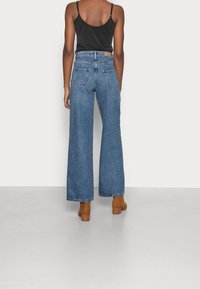 Jeans in denim blu a gamba larga con vita alta, due tasche posteriori e orli rifiniti, indossati con un top nero senza maniche e scarpe con tacco a blocco marroni.