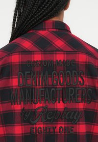 Rød og svart rutete flanellskjorte med brodert tekst på baksiden: "CUSTOM MADE DENIM GOODS MANUFACTURERS av Replay EIGHTY ONE."