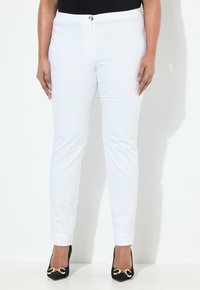 Bílé slim-fit kalhoty s hladkým povrchem, s předním zapínáním na knoflík a jemným leskem, kombinované s černými podpatky se zlatými akcenty.