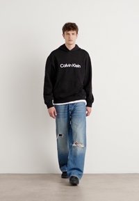 Juodas gobtuvas su baltu "Calvin Klein" logotipu, derinamas su šviesiai mėlynomis apsitempusiomis pločio kelnėmis. Modelis vilki juodus batus. Paprasta fono.