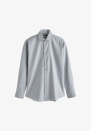 Młody mężczyzna stojący w pomieszczeniu, ubrany w lekką kraciastą koszulę na guziki, czarne spodnie i czarne eleganckie buty na tle beżowej ściany.