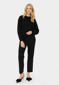 Schwarzer Strickpullover mit metallischen Sprenkeln, lockere Passform, lange Ärmel. Kombiniert mit maßgeschneiderten schwarzen Hosen und schwarzen spitzen Mules mit Schleifen.