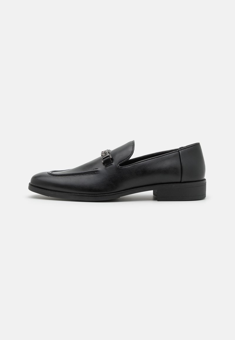 Mocassin homme en cuir noir avec un petit talon et un détail décoratif en métal en forme de mors de cheval sur le dessus, présenté sur un fond blanc uni.