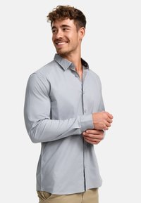 Camisa de manga larga color gris claro con cuello abotonado, con una textura suave, corte recto y pequeñas aberturas sutiles en el dobladillo.