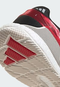 Černá sportovní obuv s červenými akcenty, která ukazuje texturovanou bílou podrážku a tři červené pruhy na patě. Zaměřte se na vzor podrážky a materiály.