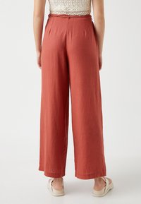 Pantalones de pierna ancha en un color óxido, confeccionados con una tela texturizada. Presentan una cintura alta con pliegues y un dobladillo limpio.