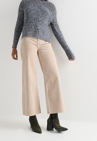 Grauer Strickpullover, hellbeige weit geschnittene Hosen und dunkelgrüne Stiefeletten mit Absatz. Texturiertes Gewebe und lockere Passform mit sichtbaren Nähten.