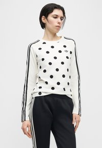 Isik, kes kannab valget pika varrukaga särki mustade täppide ja triipudega varrukatel ning musti pükse valgete küljeääriste triipudega.