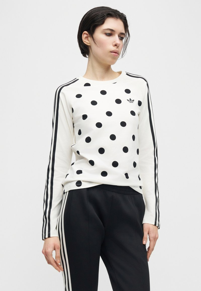 Person iført en hvid langærmet skjorte med sorte polkaprikkker og striber på ærmerne, kombineret med sorte bukser med hvide sidestipper.